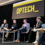 Six panelists sit on stage under an “OPTECH” sign, discussing the latest in Technology News at a conference. A moderator is seated at the left with a microphone.