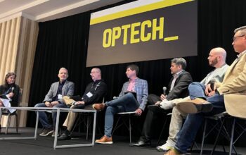 Six panelists sit on stage under an “OPTECH” sign, discussing the latest in Technology News at a conference. A moderator is seated at the left with a microphone.