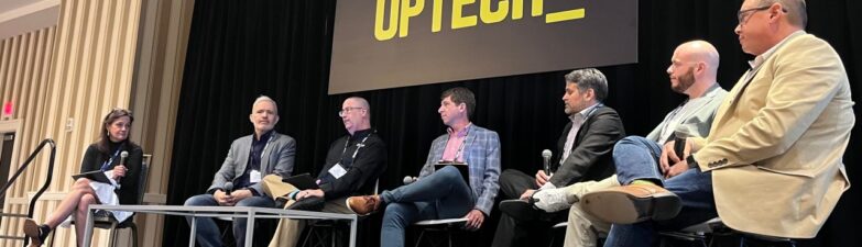 Six panelists sit on stage under an “OPTECH” sign, discussing the latest in Technology News at a conference. A moderator is seated at the left with a microphone.