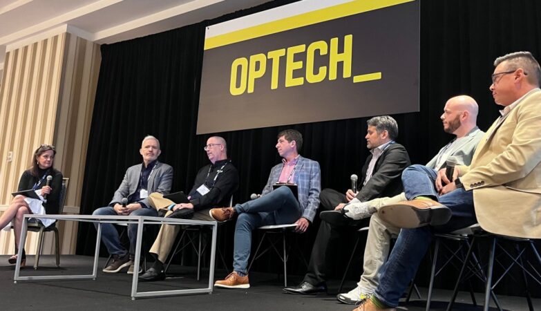 Six panelists sit on stage under an “OPTECH” sign, discussing the latest in Technology News at a conference. A moderator is seated at the left with a microphone.