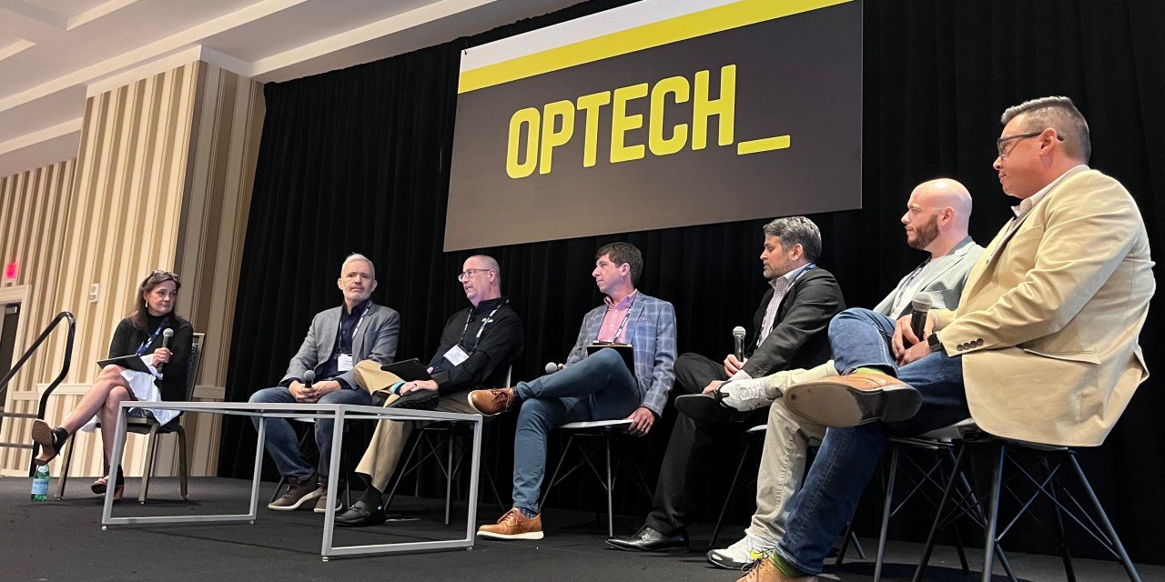 Six panelists sit on stage under an “OPTECH” sign, discussing the latest in Technology News at a conference. A moderator is seated at the left with a microphone.